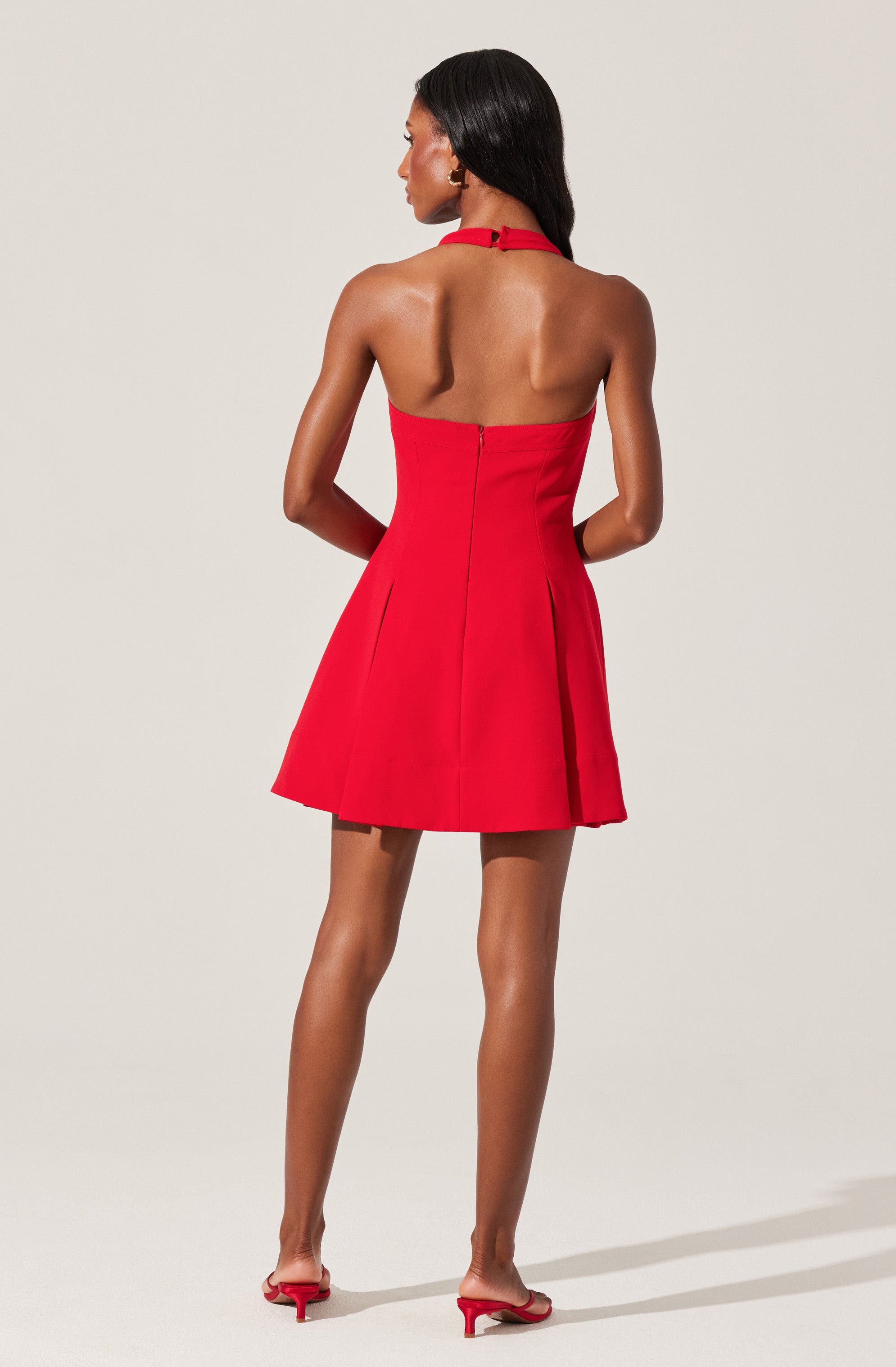 A woman is wearing a sleeveless, red, halter-neck dress with a fitted bodice and a flared, pleated skirt that falls above the knees. She has black hair styled straight and is wearing matching red open-toe heels.
