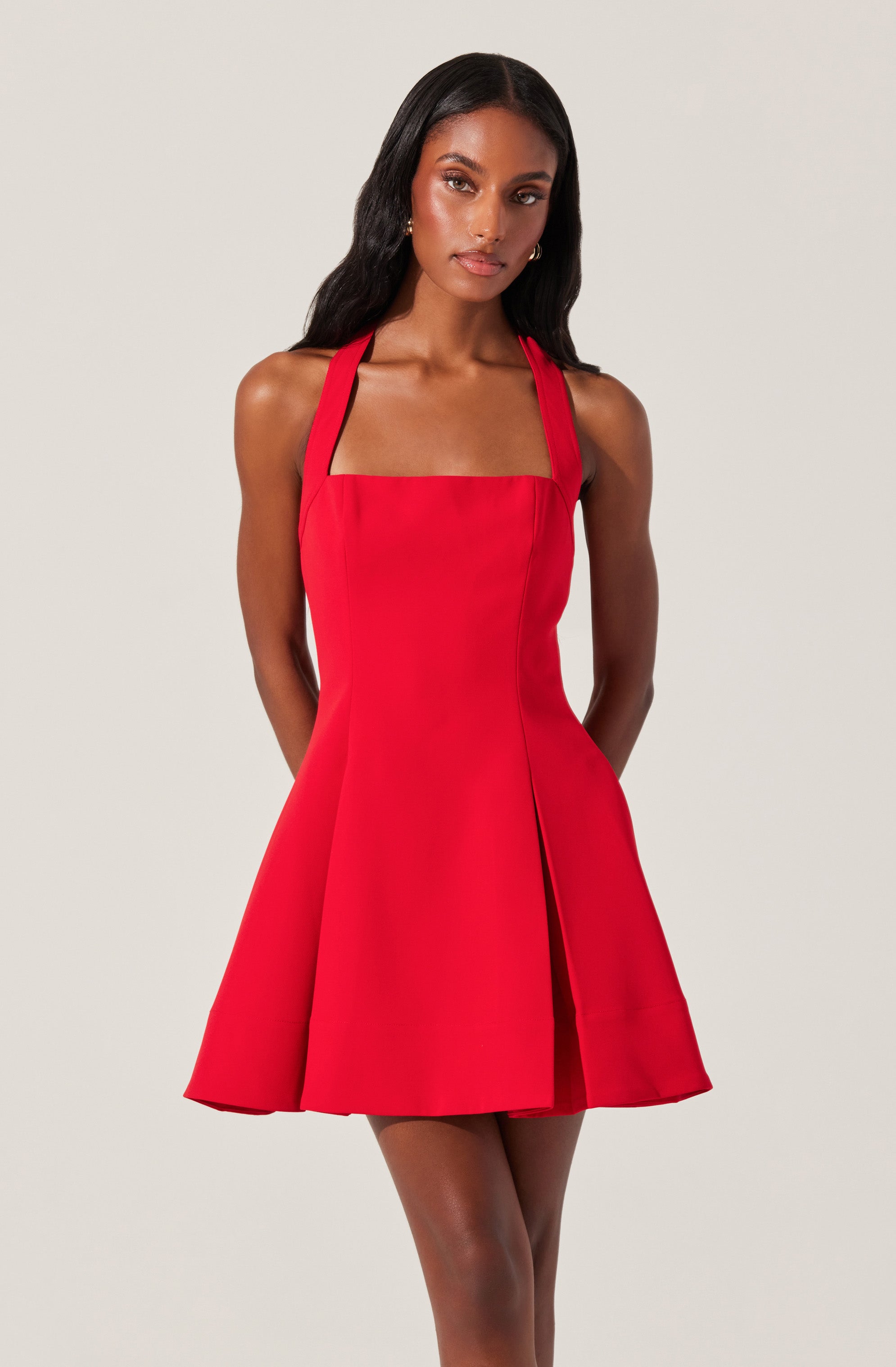 A woman is wearing a sleeveless, bright red A-line dress with a fitted bodice and a square neckline. The dress features wide straps that go over her shoulders and a front slit that adds a touch of style. The fabric appears structured, giving the dress a polished look.