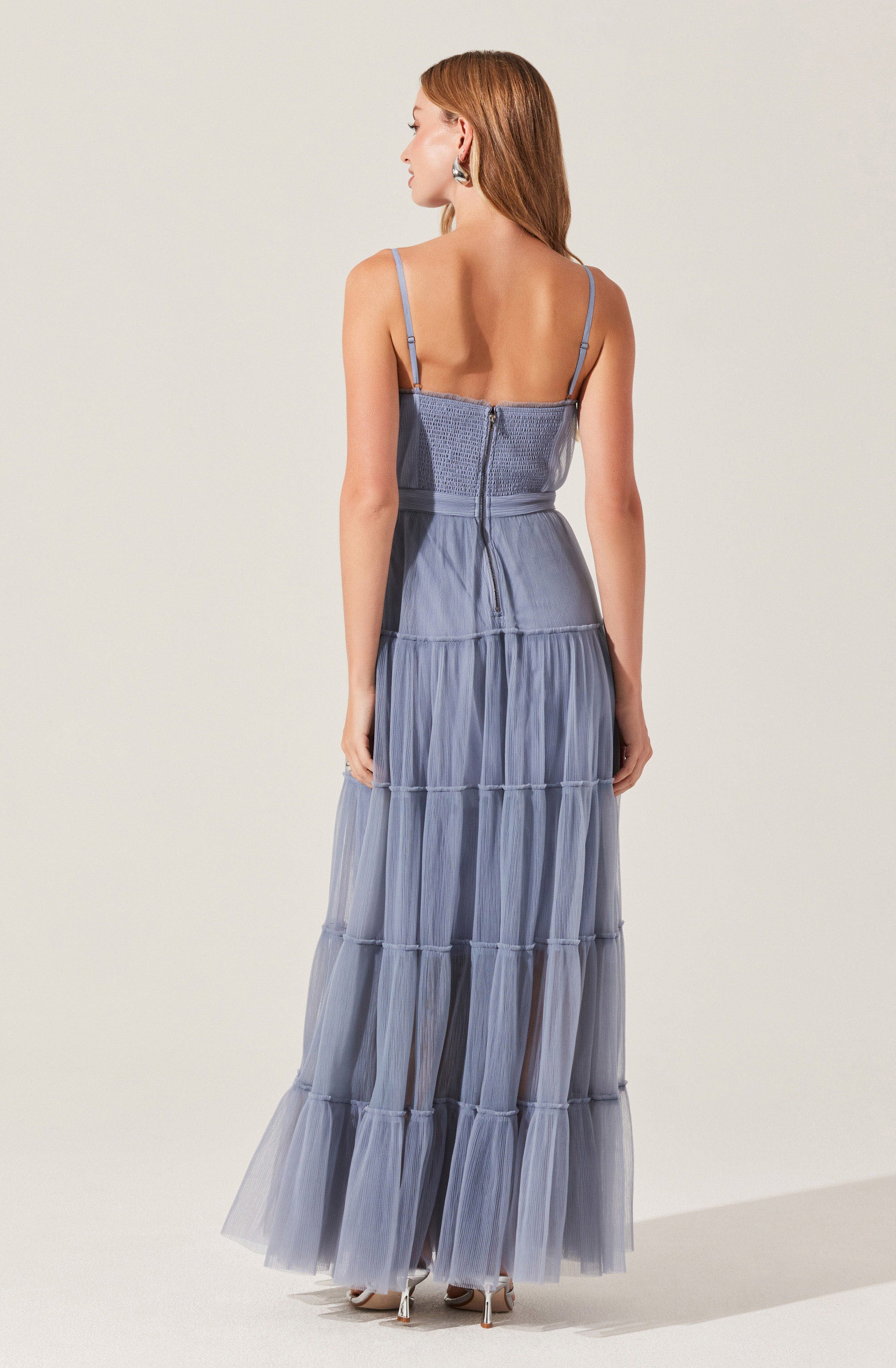A woman is wearing a long, flowing dress with thin spaghetti straps and a fitted bodice. The dress features a tiered skirt made of sheer, lightweight fabric in a soft blue-gray color. The back of the dress has a zipper closure and a drawstring detail at the waist.
