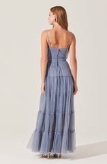 A woman is wearing a long, flowing dress with thin spaghetti straps and a fitted bodice. The dress features a tiered skirt made of sheer, lightweight fabric in a soft blue-gray color. The back of the dress has a zipper closure and a drawstring detail at the waist.