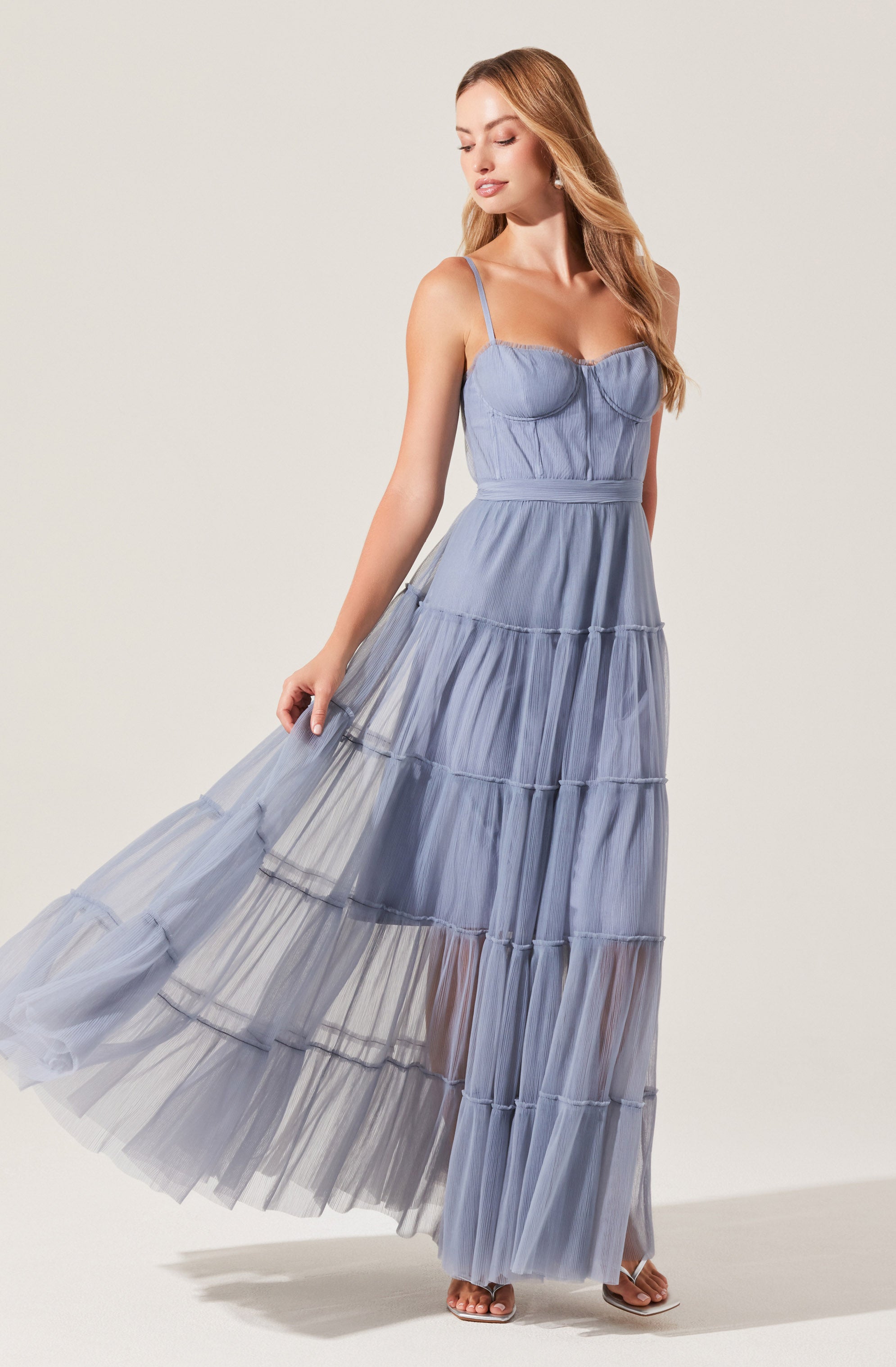 A woman is wearing a sleeveless, light blue maxi dress with thin spaghetti straps. The dress features a fitted bodice with a sweetheart neckline and vertical seam details. The skirt is made of multiple horizontal tiers of sheer, flowy fabric, creating a layered and airy appearance. She is also wearing open-toe heels.