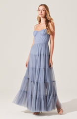 The woman is wearing a light blue, sleeveless maxi dress with spaghetti straps. The dress has a fitted bodice with a sweetheart neckline and a tiered, ruffled skirt made of a sheer, textured fabric. She is also wearing open-toe sandals and has long, wavy hair.