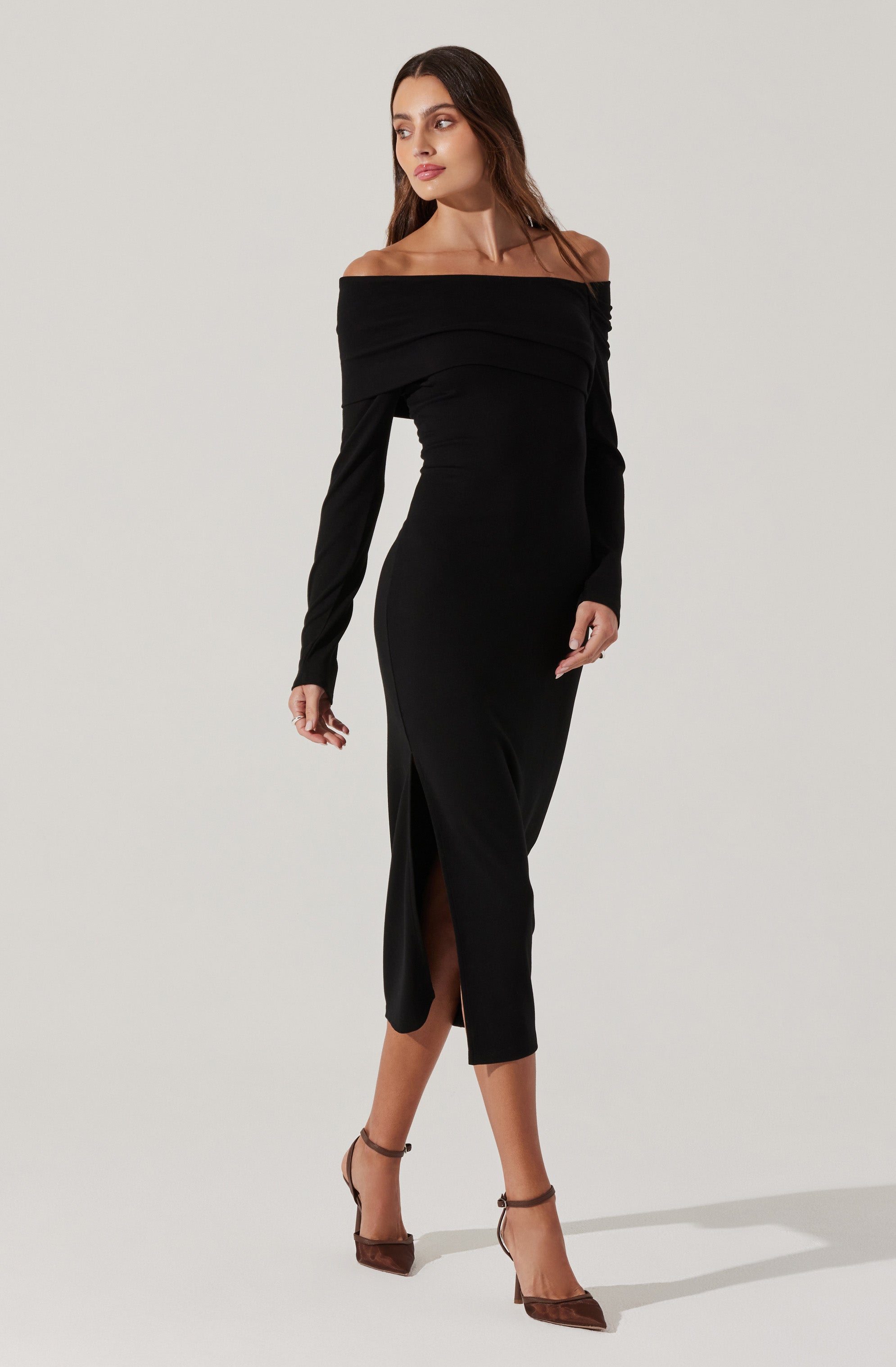 A woman is wearing a black off-the-shoulder dress with long sleeves. The dress features a fitted silhouette, horizontal pleats across the bust, and a thigh-high slit on the left side. She is also wearing brown high-heeled shoes with ankle straps.