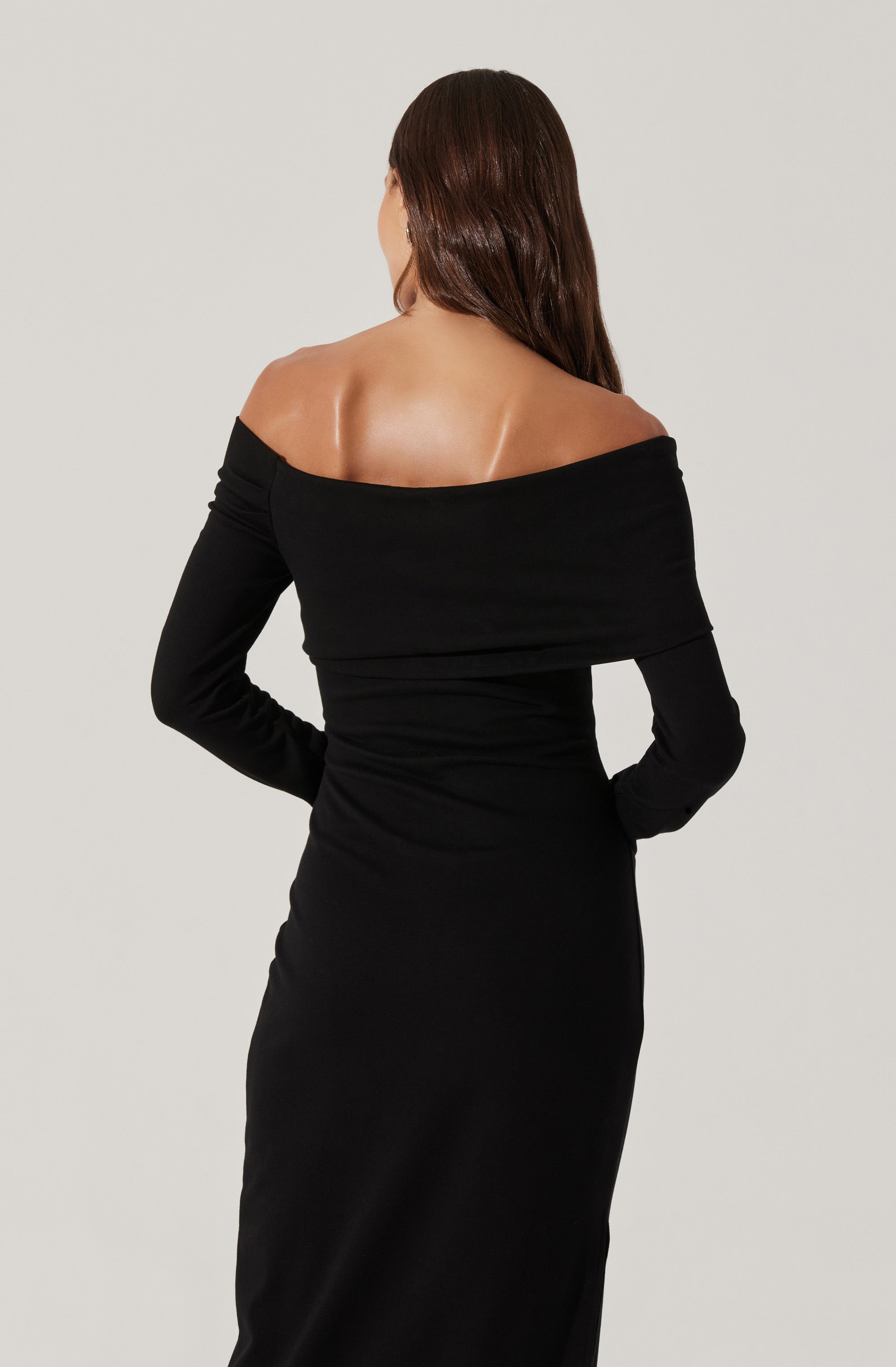 A woman is wearing an off-the-shoulder black dress with long sleeves. The dress is fitted, reaching down to her ankles.