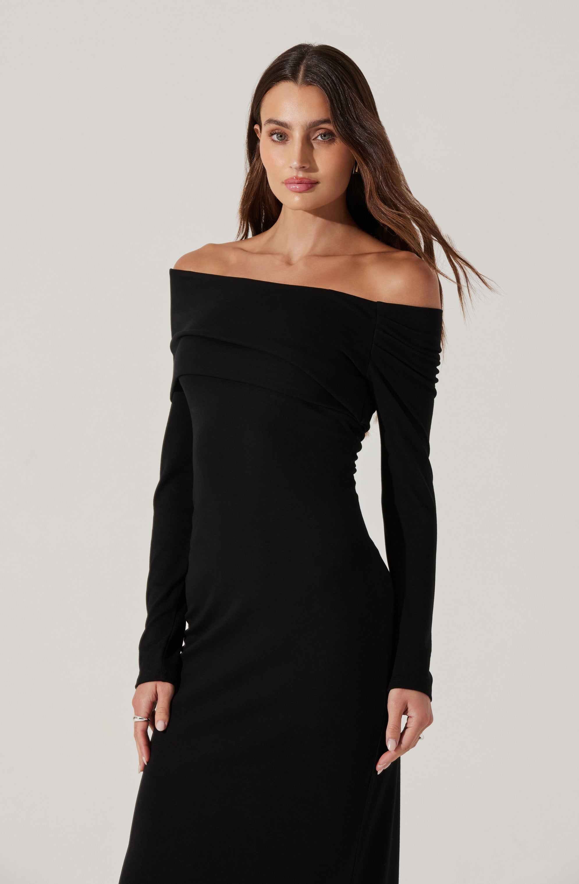 A woman is wearing a fitted, long-sleeved black dress with an off-the-shoulder neckline. The dress features a layered, draped design across the chest area.