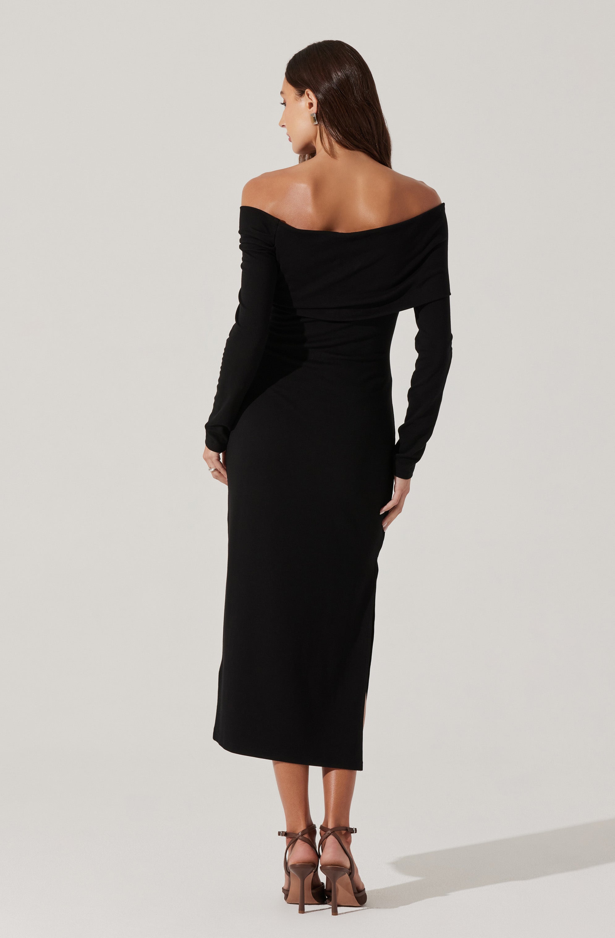 A woman is wearing an elegant off-the-shoulder black dress with long sleeves. The dress is fitted and extends to her ankles. She is also wearing high-heeled sandals with ankle straps.