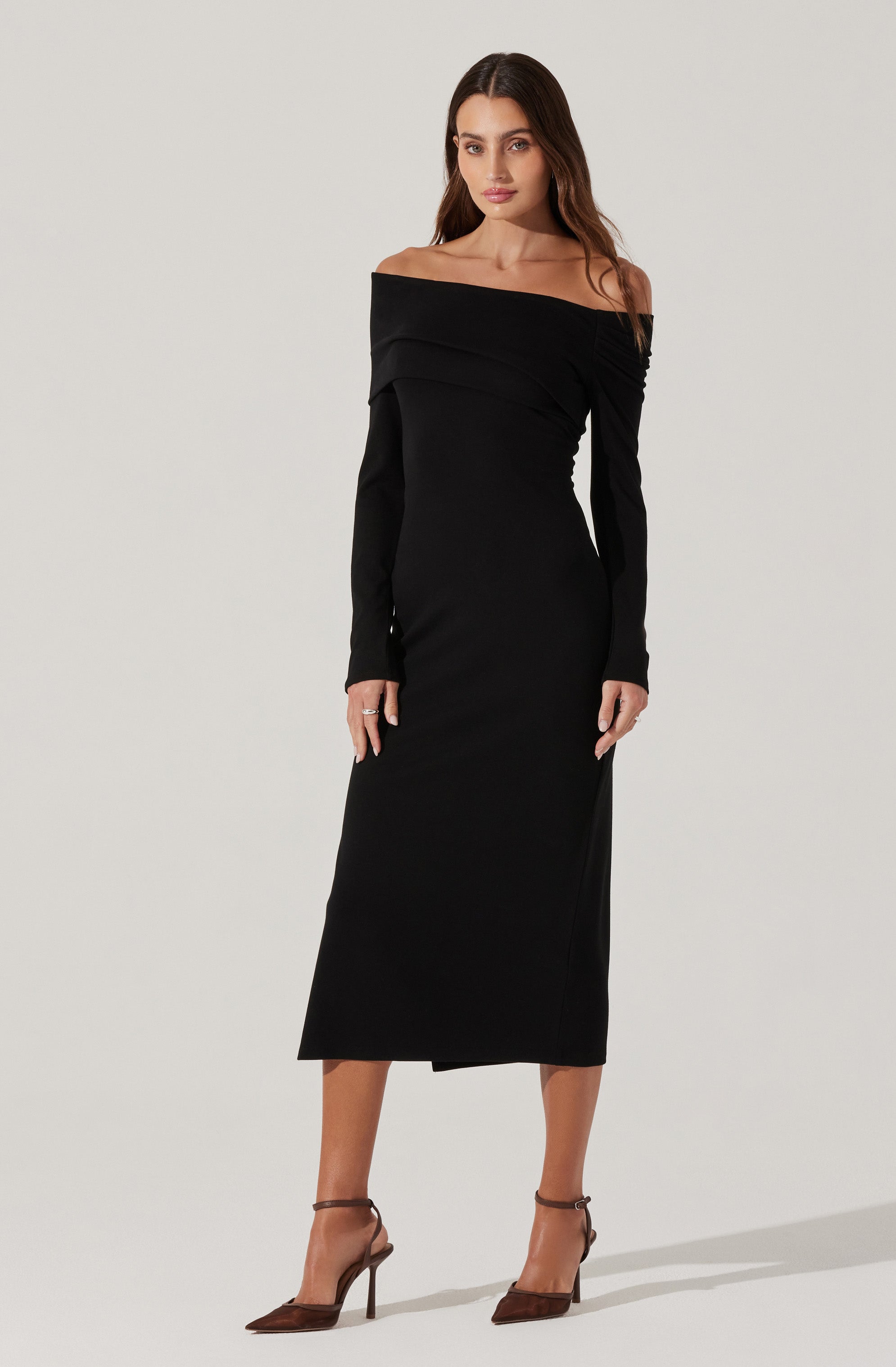 A woman is wearing a long, black, off-the-shoulder dress with long sleeves. The dress has a fitted silhouette that extends to mid-calf length. She is also wearing a pair of brown strappy high-heeled shoes.