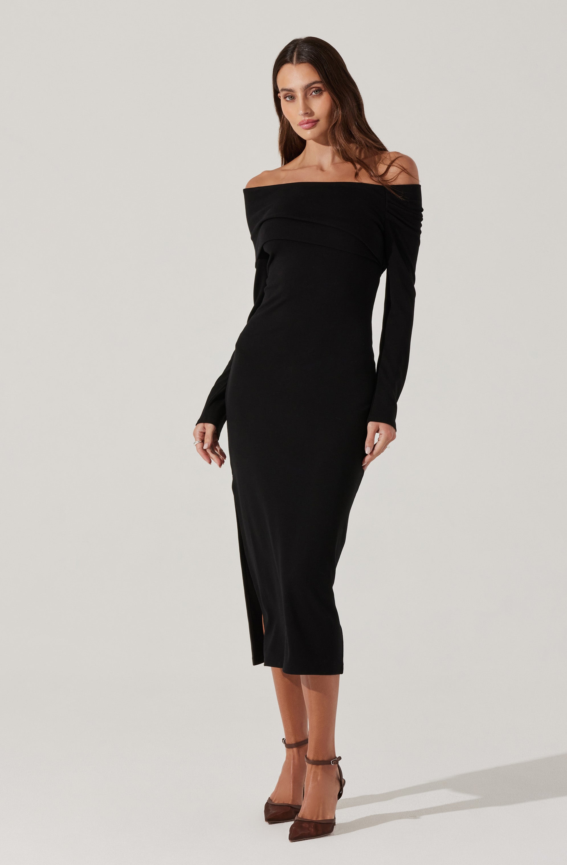 A woman is wearing a long, off-the-shoulder black dress with long sleeves. The dress fits snugly and extends to her mid-calf, featuring a subtle slit on the side. She is also wearing brown high-heeled shoes with ankle straps.