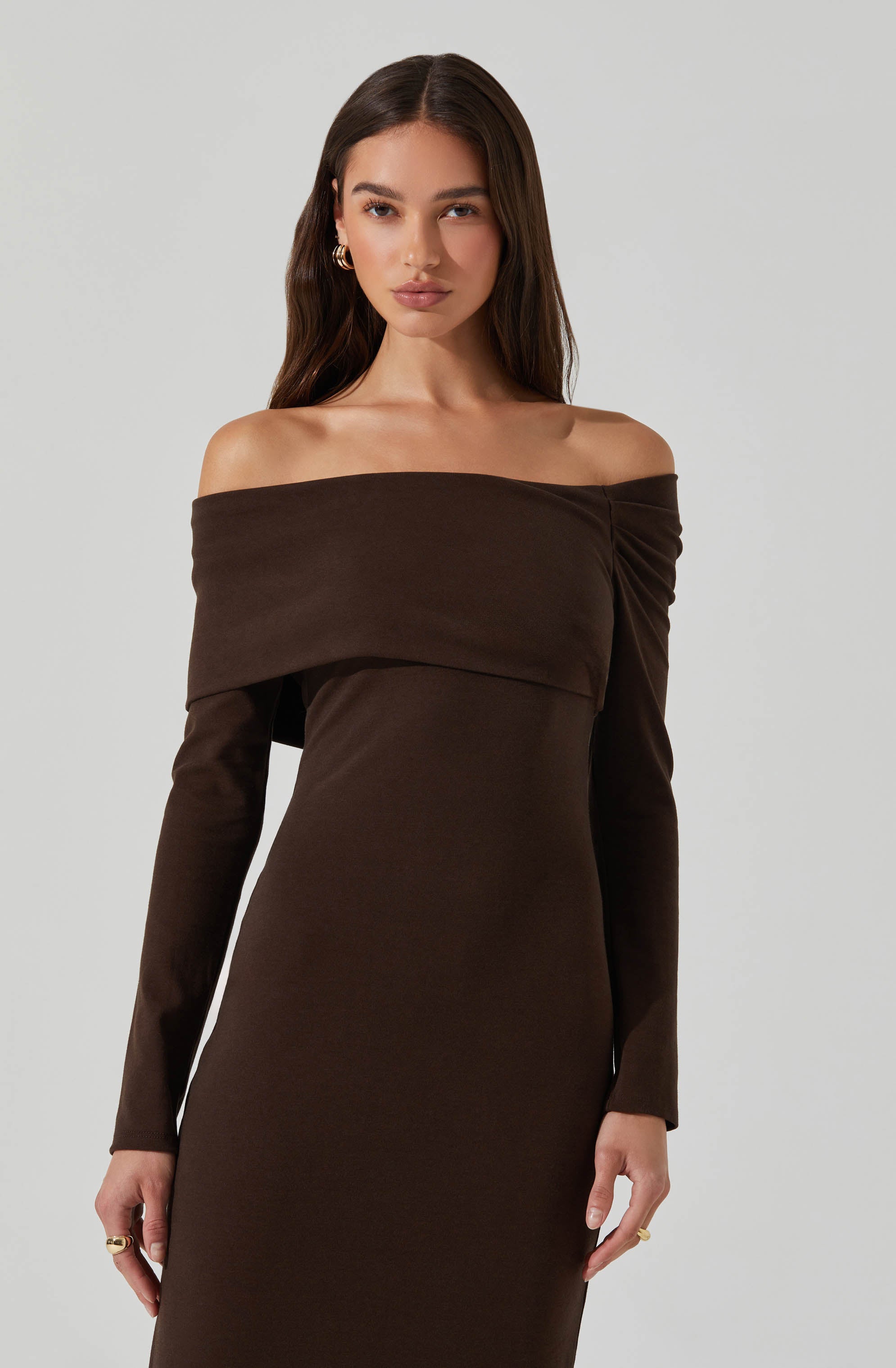 A woman wearing an off-the-shoulder, long-sleeved dress in a rich, dark brown color. The dress features a layered, wide neckline that drapes over her shoulders and upper arms, creating a sophisticated and stylish look. The fabric appears smooth and slightly stretchy, fitting her figure closely.