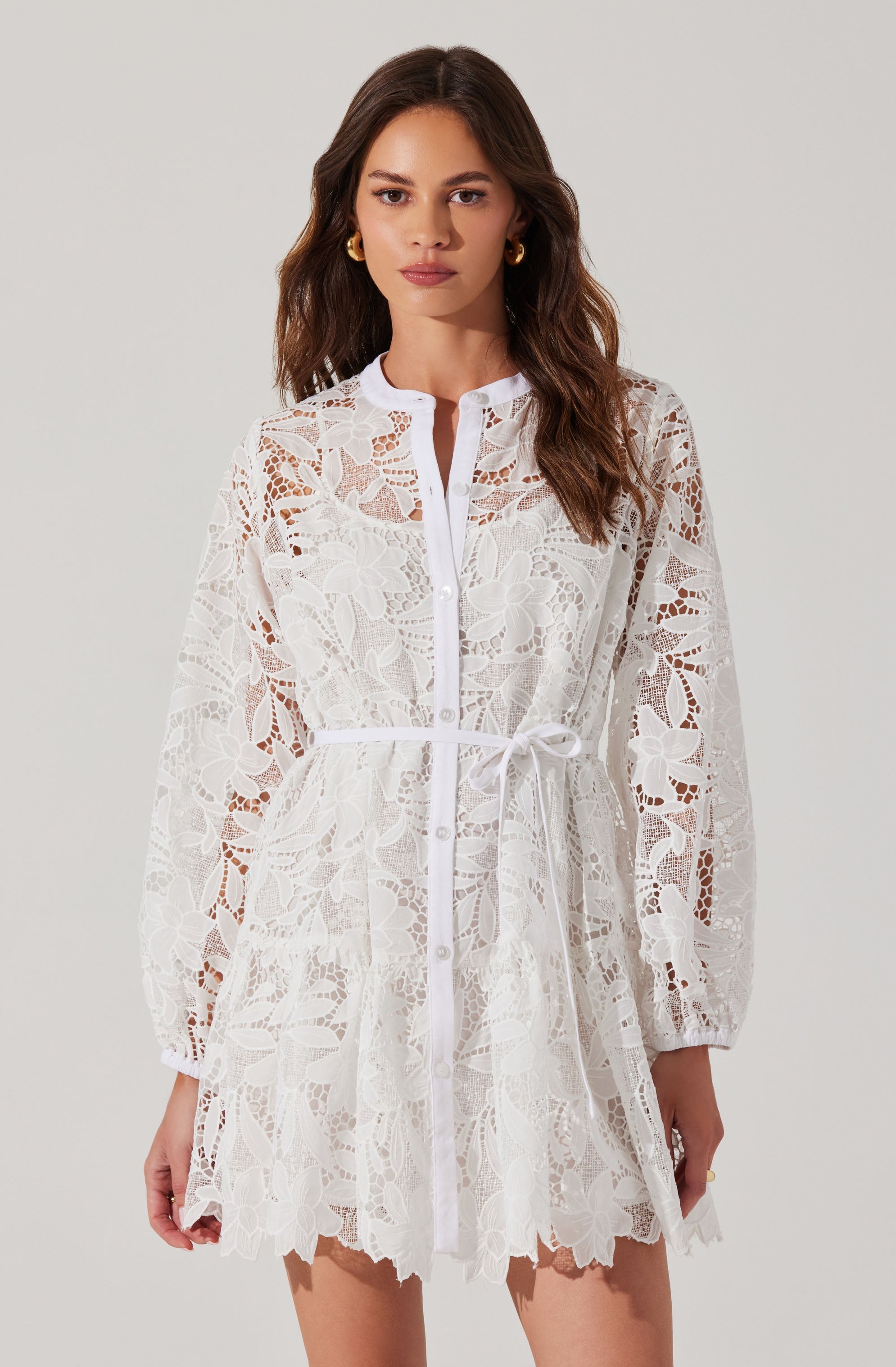 A woman is wearing a white lace dress with a floral pattern. The dress features long, sheer sleeves with elastic cuffs, a button-down front, and a waist tie. The hem of the dress is scalloped, and the lace fabric adds a delicate, intricate detail to the garment.