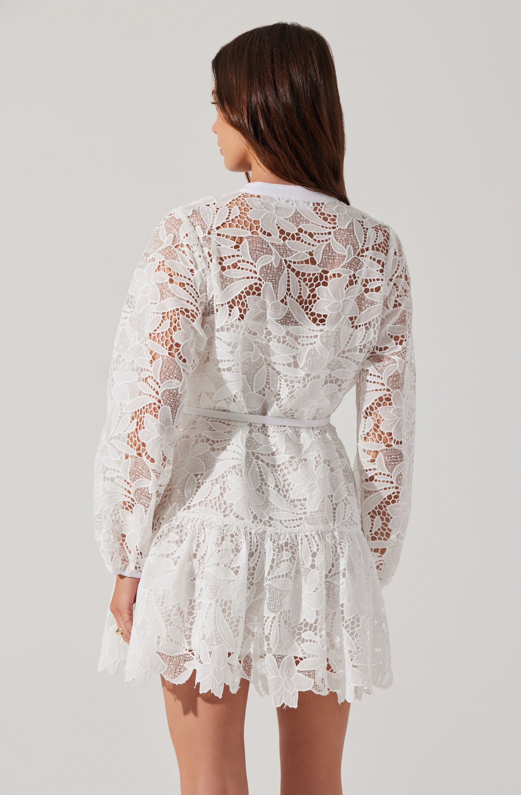 A woman is wearing a white lace dress with an intricate floral pattern. The dress features long, sheer sleeves and a tiered, ruffled skirt with scalloped edges. The lace fabric displays large floral motifs and geometric designs throughout.
