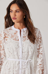 A woman is wearing a white, semi-sheer lace dress with floral patterns. The dress features a collarless neckline, button-down front, and a matching fabric belt tied at the waist.