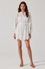 A woman is wearing a white lace dress with long sleeves and a high neckline. The dress features intricate lace patterns with floral and geometric designs, a front zipper closure, and a matching waist tie that accentuates her figure. The hem of the dress has a scalloped edge, and the see-through lace adds a delicate, feminine touch. She pairs the dress with neutral-colored open-toe heels.