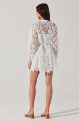 A woman is wearing a white lace dress with long, semi-sheer sleeves and a belt at the waist. The dress has a floral lace pattern, a ruffled hem, and a slightly fitted silhouette. She is also wearing beige open-toe sandals with a small heel.