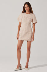 The dress is a short, cream-colored, textured fabric with a slightly loose fit. It has a round neckline and short, wide sleeves. The hem of the dress falls midway between the hips and knees. The model is wearing white high-heeled shoes with ankle straps and gold earrings.