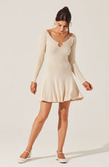 A woman wearing a cream-colored, long-sleeved knit dress with a ribbed texture. The dress has a keyhole opening at the neckline with a small metal ring detail. The skirt portion of the dress is flared and falls above the knees. She is also wearing white heels.