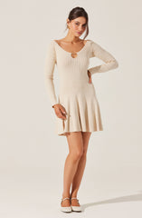 A woman is wearing a cream-colored, long-sleeve knit sweater with a round neckline and a small keyhole cutout at the front. She pairs it with a matching cream-colored pleated skirt that falls above the knees. She completes the look with white heeled shoes.