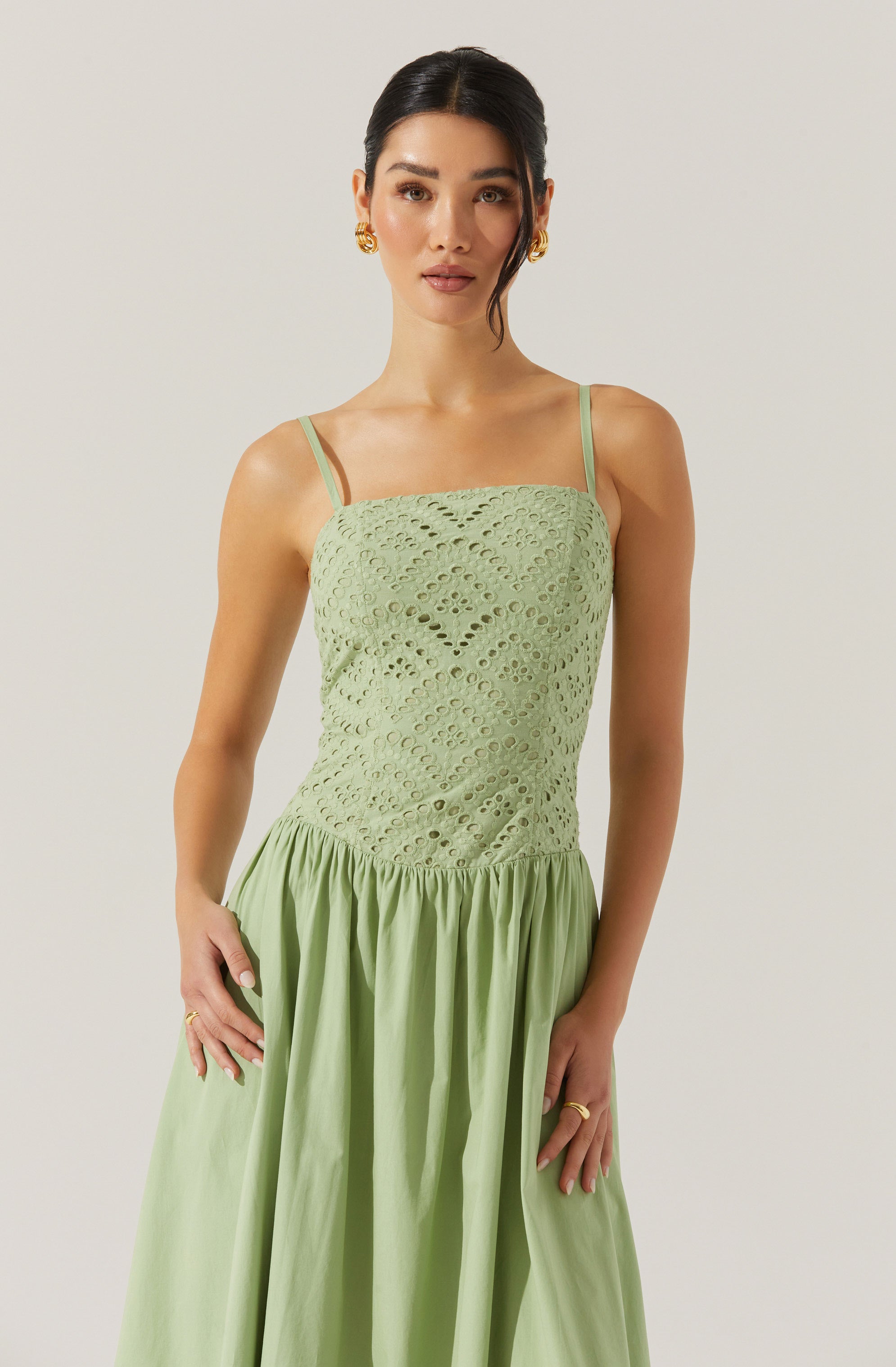 A woman is wearing a light green dress with thin spaghetti straps. The top portion of the dress features a textured, eyelet fabric with circular and diamond-shaped cutouts. The skirt of the dress is gathered at the waist, flowing freely with a smooth, solid fabric. She has dark hair styled in a sleek updo, and she is accessorizing with gold earrings and rings.