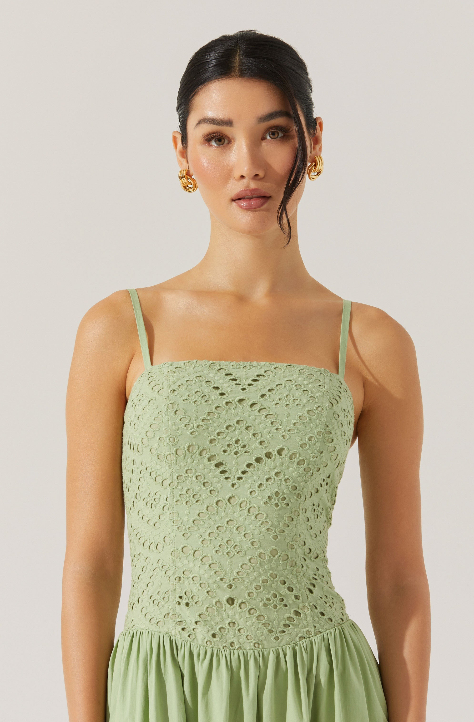 A woman is wearing a light green, sleeveless dress with thin spaghetti straps. The top portion of the dress features a textured, eyelet embroidery pattern with geometric and floral shapes, creating a lace-like appearance. The skirt of the dress is gathered and flows gently from the waist, made of a smooth, lightweight fabric.