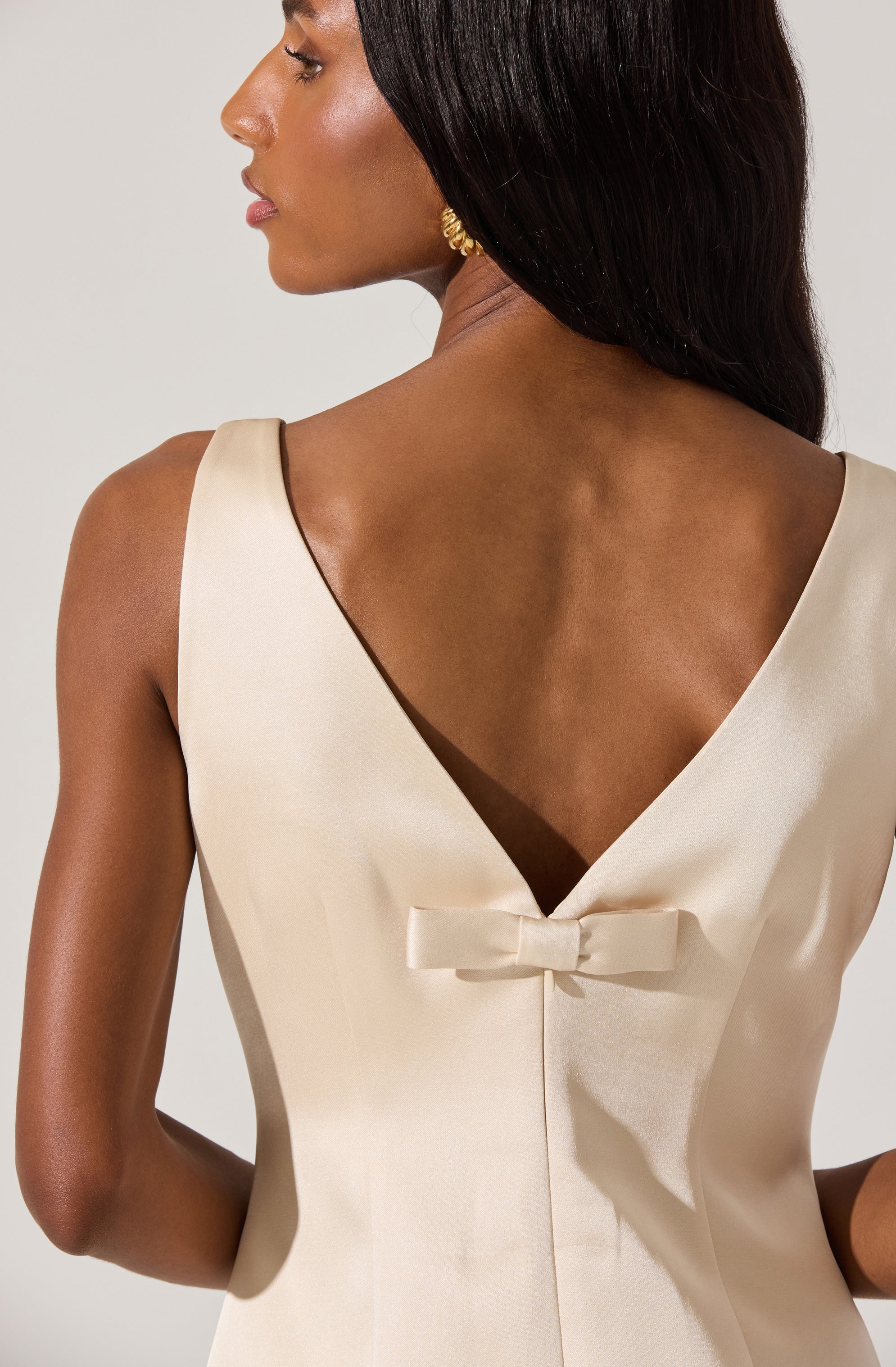 A woman is wearing a sleeveless, cream-colored satin dress with a V-shaped back. The dress features a small, decorative bow at the lower part of the back. She has long, dark hair and is accessorized with gold earrings.