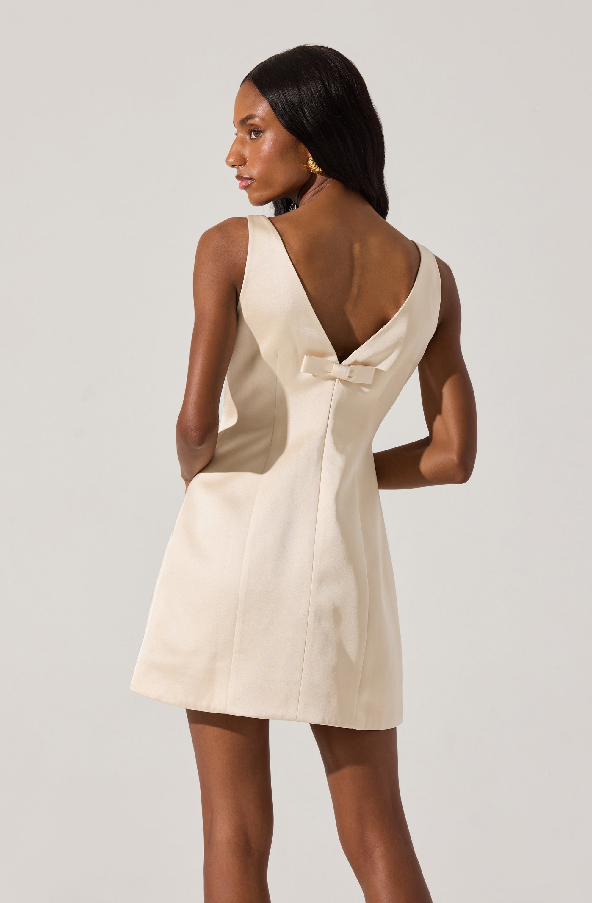 A woman wearing a sleeveless, cream-colored dress with a V-neckline at the back. The dress features a small bow detail at the center of the back near the neckline. The fabric appears smooth and structured, with vertical seams running down the front and back.
