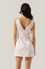 A woman is wearing a sleeveless, satin-like dress in a soft, light pink color. The dress has a deep V-back neckline with a small, decorative bow at the waist. The fabric appears smooth and shiny, giving it an elegant look.