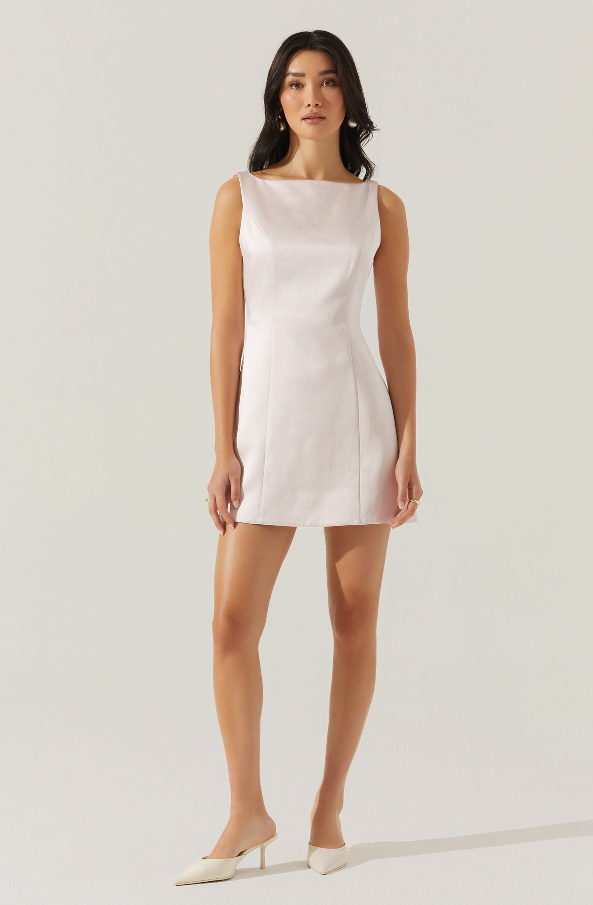 A woman is wearing a sleeveless, ivory-colored, fitted mini dress with a boat neckline and subtle vertical seam details. She pairs the dress with pointed-toe, ivory-colored high heels.