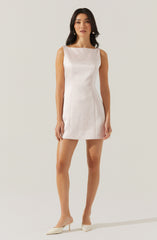 A woman is wearing a sleeveless, ivory-colored, fitted mini dress with a boat neckline and subtle vertical seam details. She pairs the dress with pointed-toe, ivory-colored high heels.