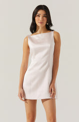 A woman is wearing a sleeveless, off-white or cream-colored satin or silk mini dress with a boat neckline. The dress has a smooth, shiny finish and fitted silhouette. She accessorizes with pearl earrings and a gold ring on her right hand.