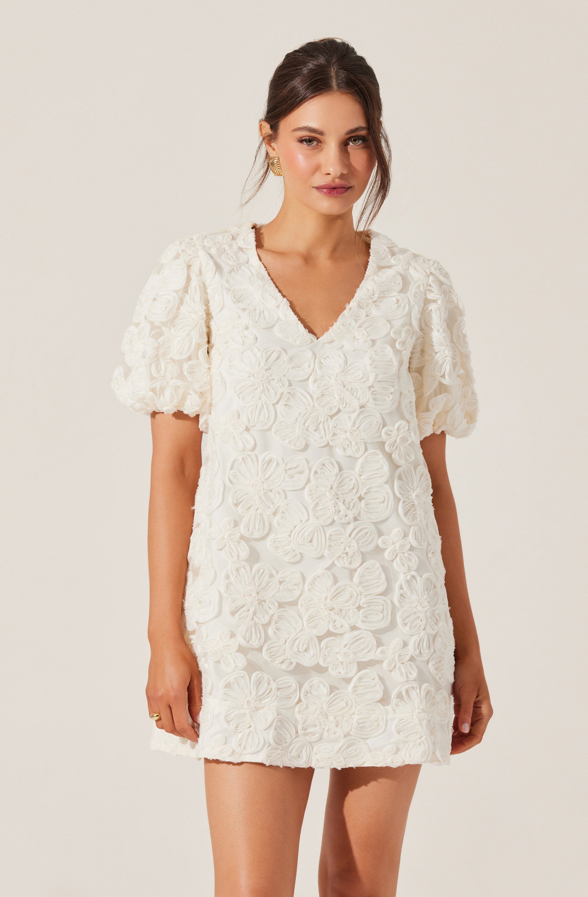 A woman is wearing a white dress with a textured floral pattern. The dress features puffed short sleeves and a V-neckline, with intricate flower motifs covering the entire garment.