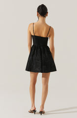 The woman is wearing a black dress with thin spaghetti straps. The dress has a fitted bodice and a full, slightly flared skirt with subtle floral patterns. She is also wearing small earrings and black heeled sandals.