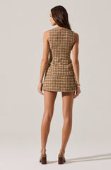 A woman is wearing a sleeveless, fitted, knee-length dress with a tartan plaid pattern in shades of brown, beige, and black. She is also wearing high-heeled shoes with a beige upper and dark brown block heel.