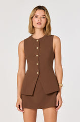 A woman is wearing a sleeveless, fitted brown dress with a matching blazer-like top that features a high, round neckline. The top has a row of gold buttons down the front, and the blazer's hem flares slightly at the waist. The matching skirt is a straight, above-the-knee length. She is accessorized with gold bracelets on her right wrist and a gold ring on her left hand.