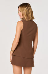 A woman is wearing a sleeveless, fitted brown dress with a peplum-style hem at the waist. The dress is made of a smooth fabric with a slight sheen. She has shoulder-length, wavy blonde hair and is accessorizing with gold bangles on her left wrist and a ring on her left hand.