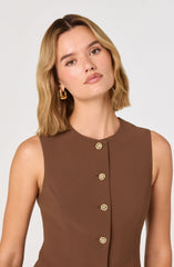 A woman with shoulder-length wavy blonde hair is wearing a sleeveless brown dress with a round neckline. The dress features a row of decorative gold buttons running down the front. She has on large, gold hoop earrings.