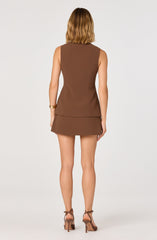 A woman is wearing a sleeveless, brown, fitted top with a peplum hem and a matching mini skirt. She has paired her outfit with strappy high-heeled sandals and is accessorizing with stacked gold bracelets on her left wrist.