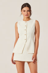 A woman is wearing a sleeveless, cream-colored tailored top with a round neckline and decorative buttons down the front. The top has a subtle peplum style with a fitted waist and an asymmetrical front opening. She is also wearing a matching cream-colored mini skirt. Her hair is styled in a loose, elegant updo, and she is accessorized with small earrings and a ring on her right hand.
