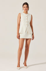 A woman is wearing a sleeveless, cream-colored suit consisting of a fitted, button-front blazer with decorative buttons and a matching mini skirt. She is also wearing cream-colored pointed-toe heels.