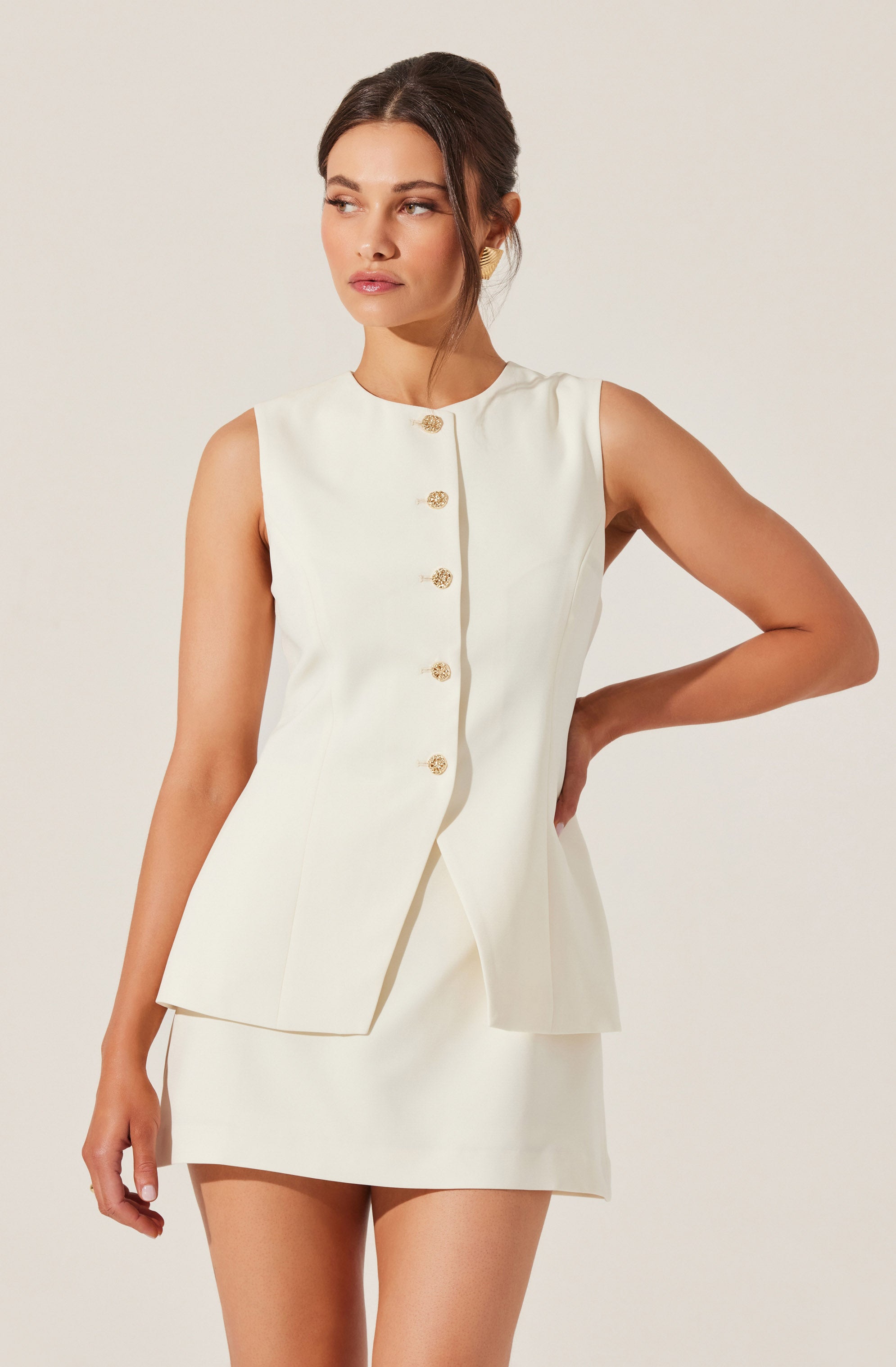 A woman is wearing a sleeveless, cream-colored suit consisting of a fitted vest with a rounded neckline and decorative gold buttons down the front, paired with a matching short skirt. The overall style is elegant and sophisticated.