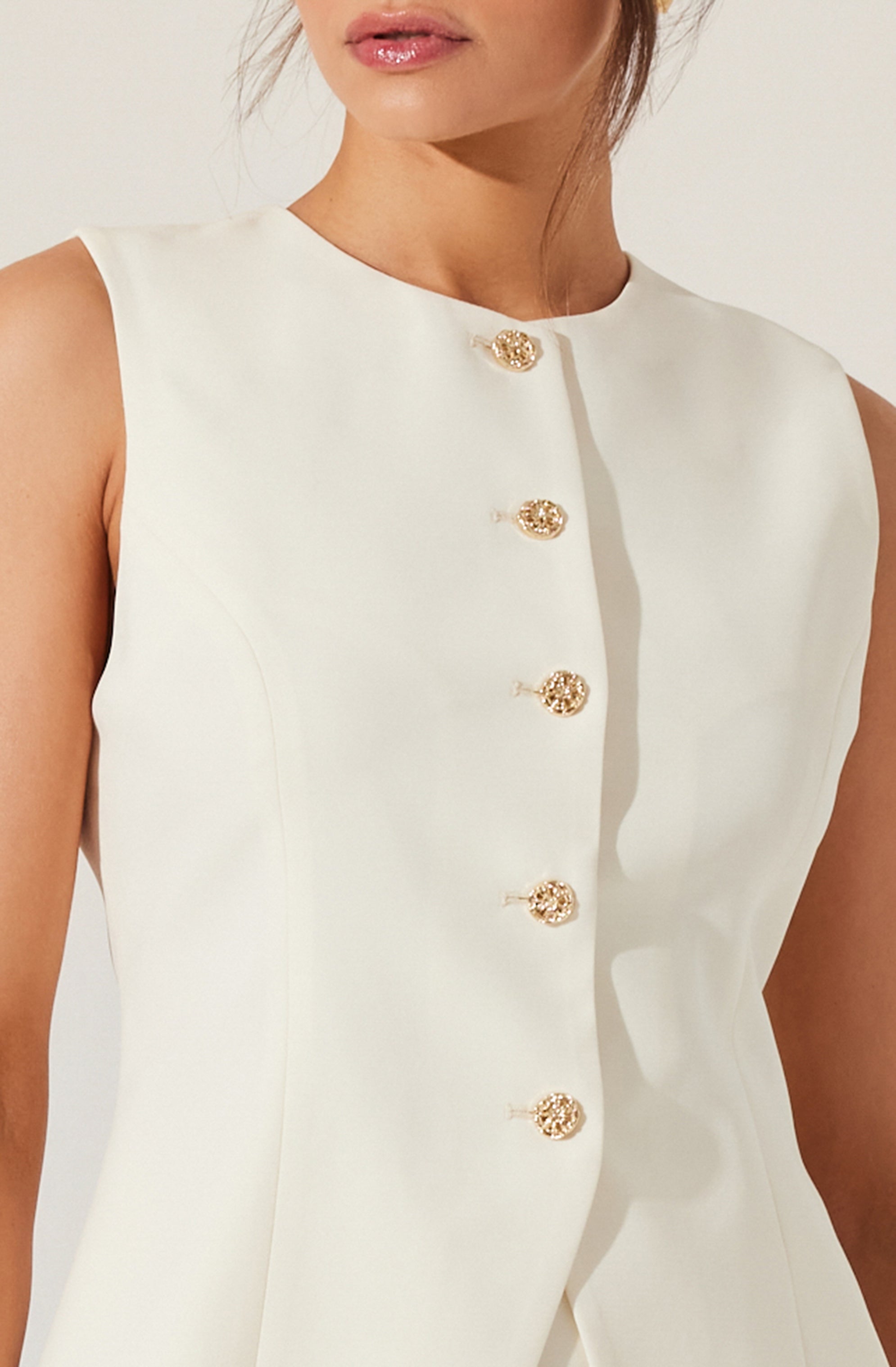 A woman wearing a sleeveless white dress with a round neckline, featuring a row of gold and pearl decorative buttons along the front.