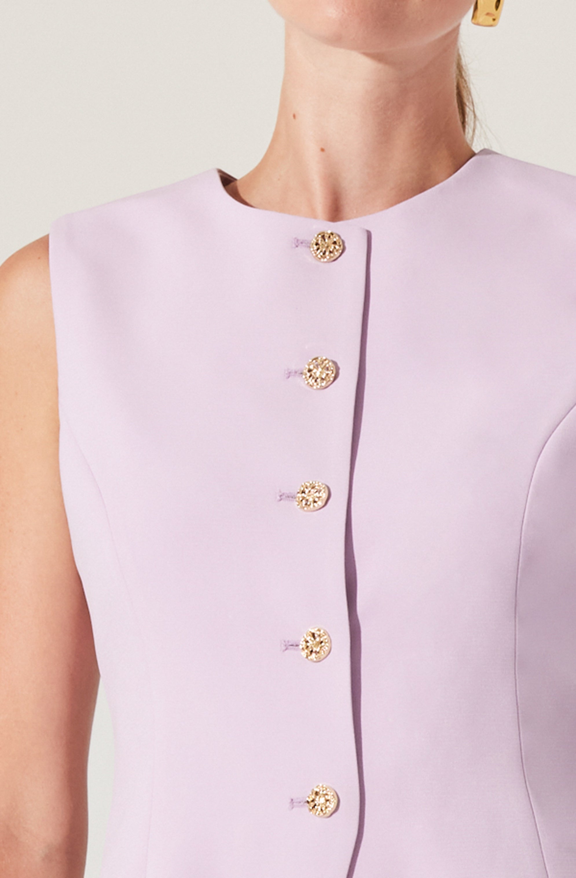 A sleeveless, light lavender-colored dress with a round neckline. The dress features a row of gold buttons down the front, with intricate detailing, and has a subtle vertical seam detail on the side.