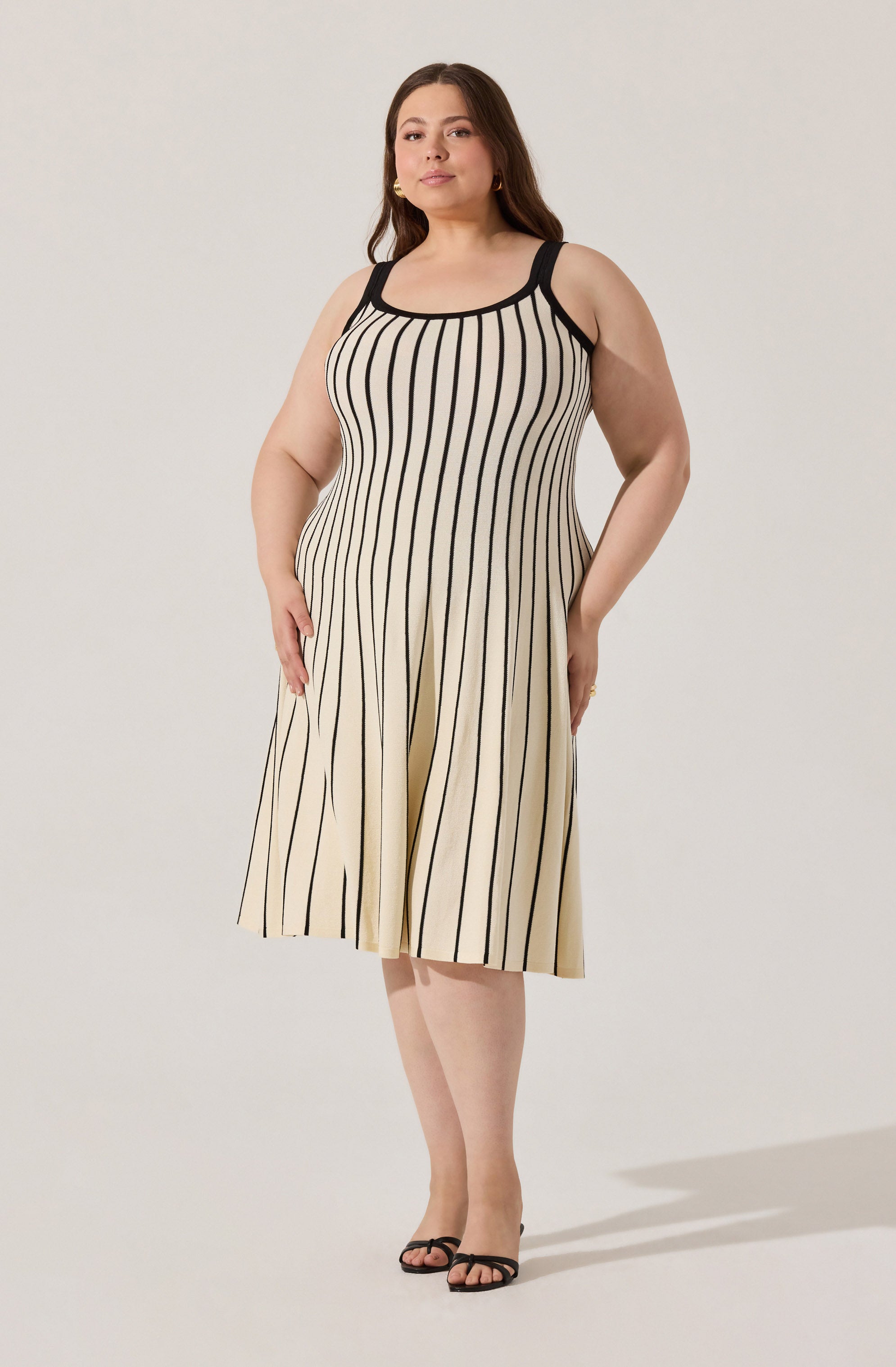 A woman is wearing a sleeveless, knee-length dress with vertical black and beige stripes. The dress has black trim around the neckline and shoulder straps. She is also wearing open-toed black heels.