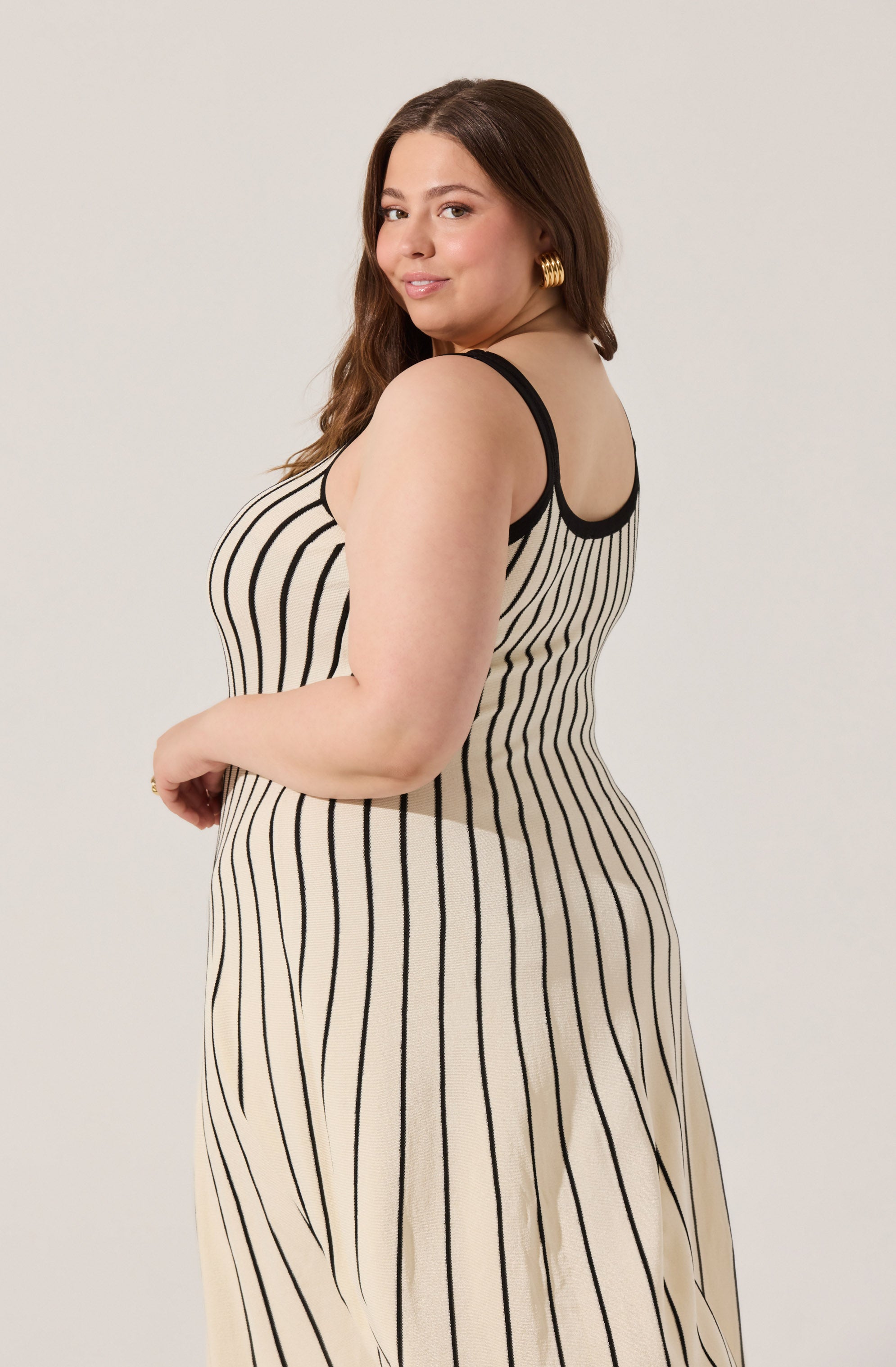 A woman is wearing a sleeveless dress with thin black straps and a fitted bodice. The dress features vertical black stripes on a beige background, creating a classic pinstripe pattern throughout.