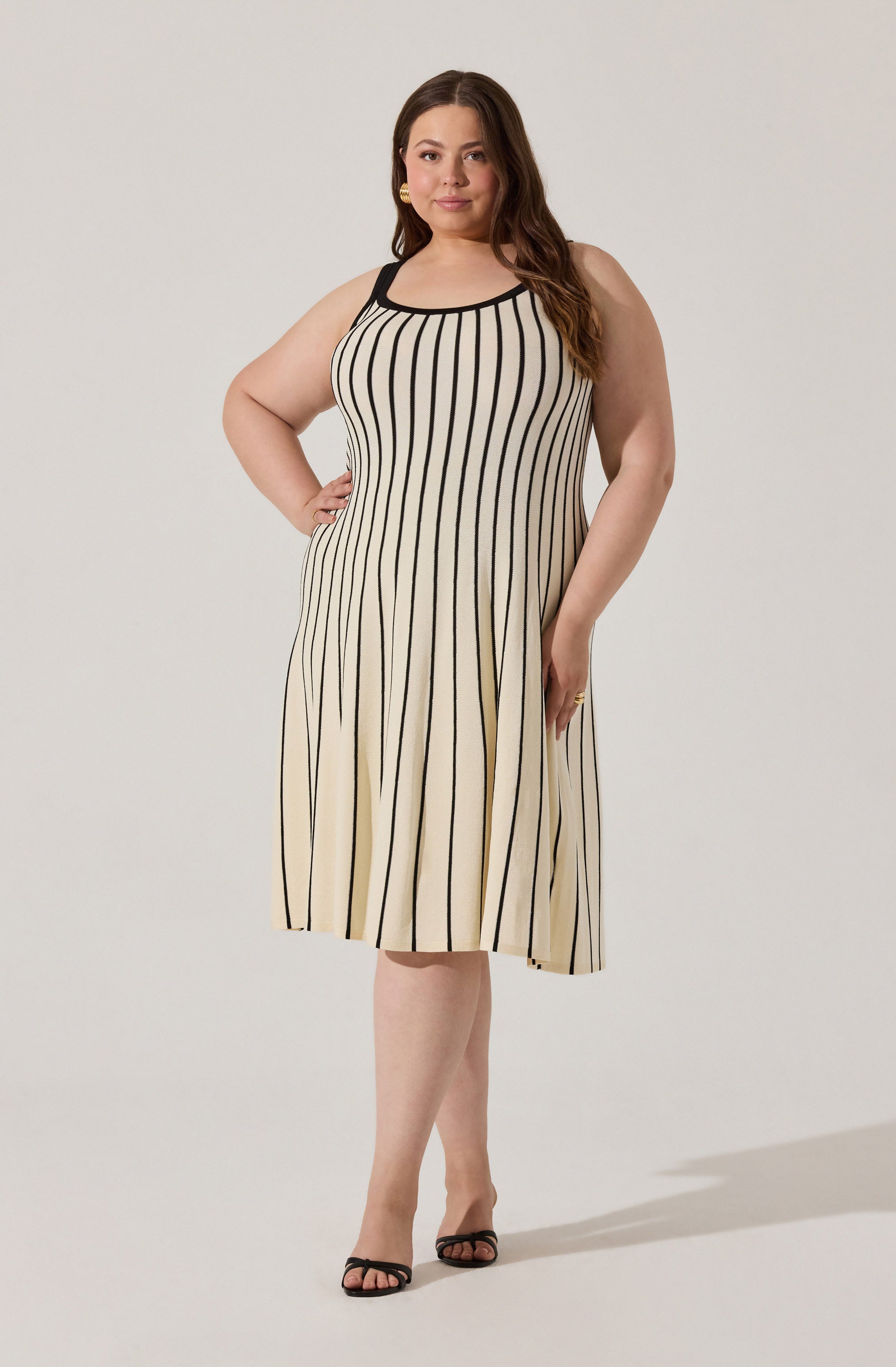 A woman is wearing a sleeveless, knee-length dress with a fitted bodice and a flared skirt. The dress features vertical black stripes on a cream background, with black trim along the neckline and armholes. She completes the look with black open-toe heels.