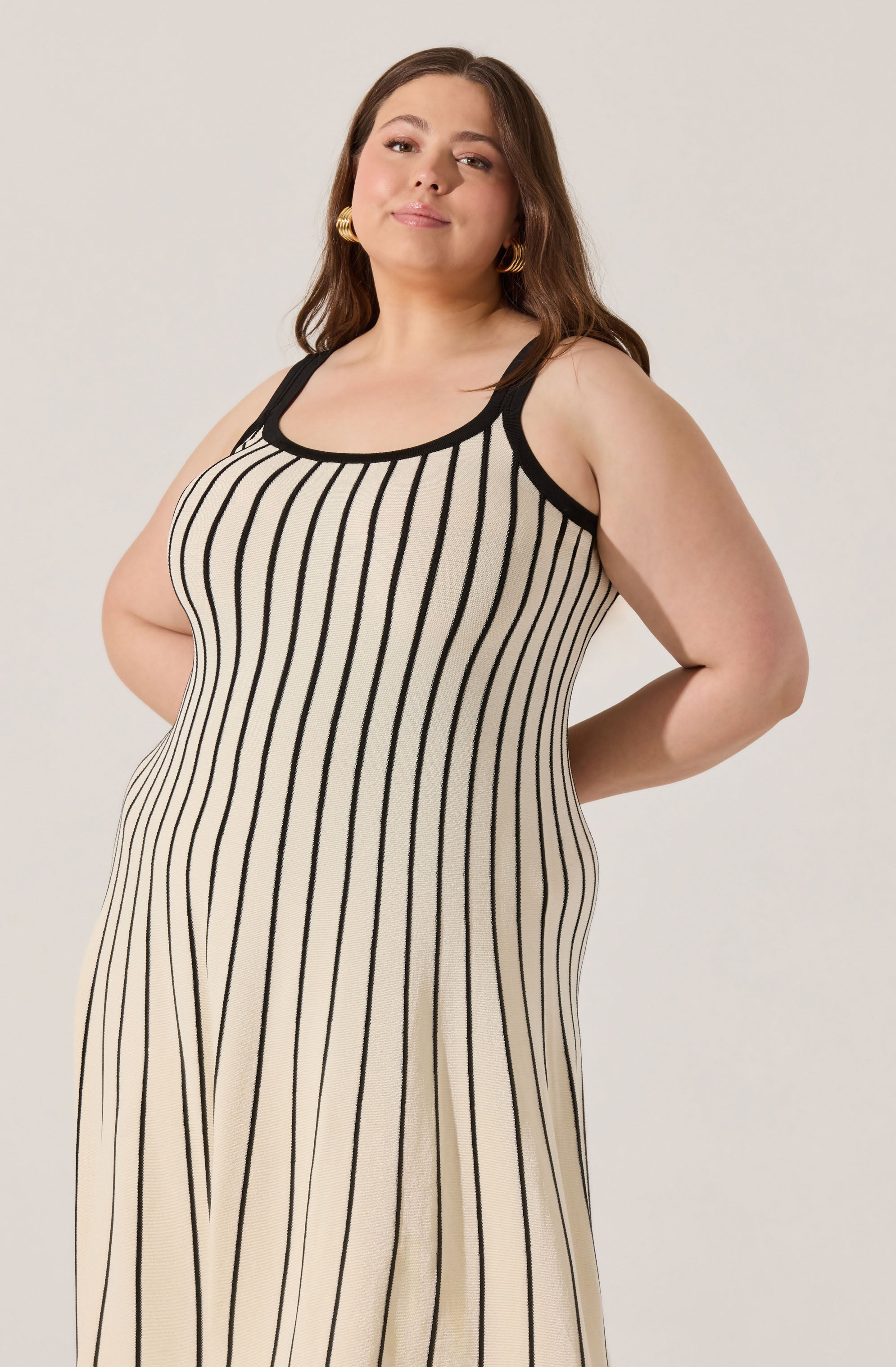 A woman is wearing a sleeveless, beige dress with vertical black stripes. The dress has a scoop neckline and black trim along the neckline and armholes.