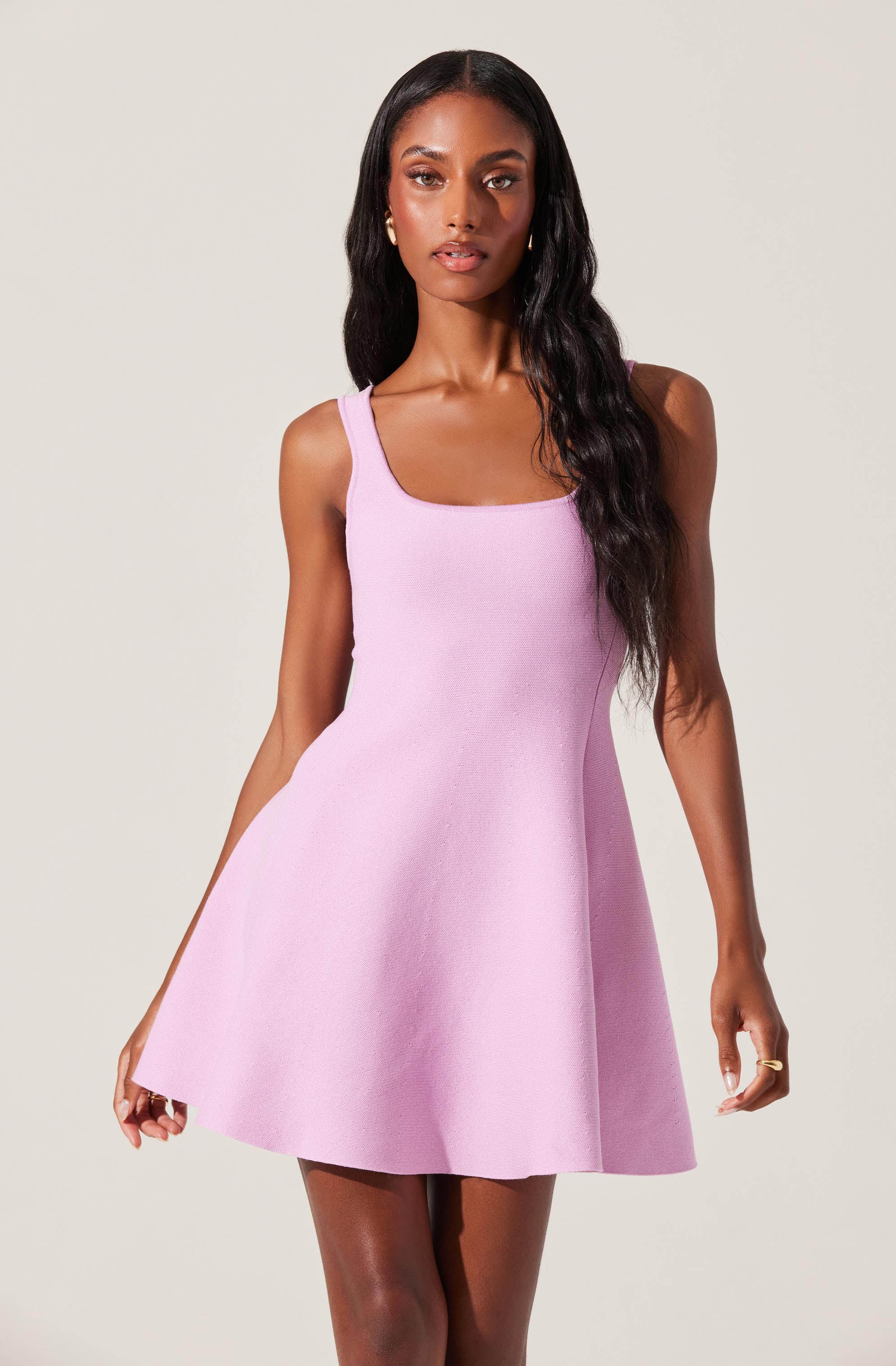A woman is wearing a sleeveless, light purple dress with a fitted bodice and a flared skirt that falls above the knees. The dress has wide shoulder straps and a square neckline.