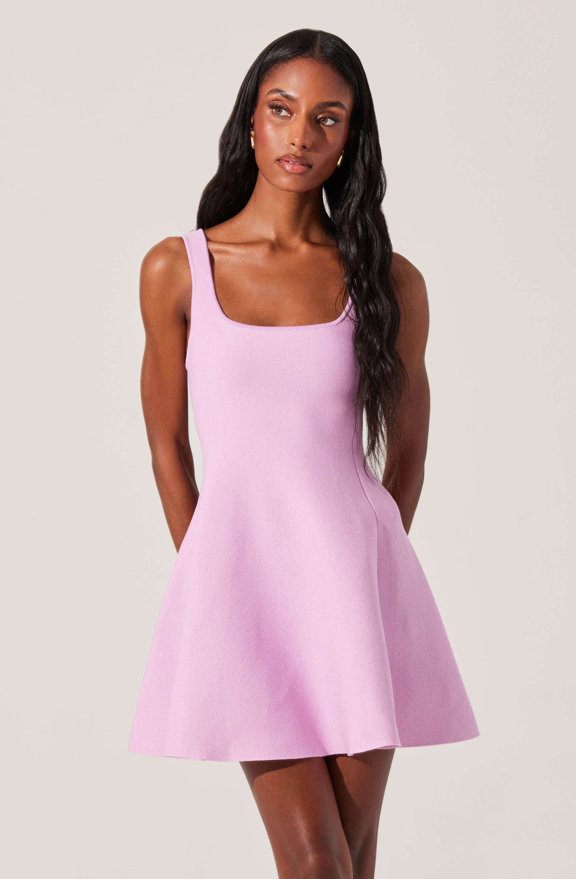 A woman is wearing a sleeveless, pastel pink dress with thick straps, a fitted bodice, and a flared skirt that falls above the knees.