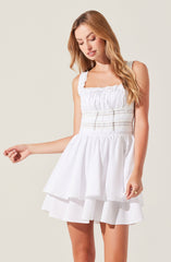 A young woman is wearing a sleeveless white dress with a square neckline, featuring ruffled detailing along the bodice and shoulder straps. The dress has a fitted waistband with decorative horizontal stitch details and a layered, flared skirt that ends above the knees.