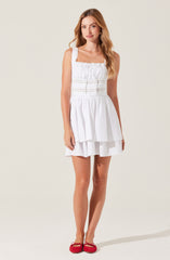 The woman is wearing a sleeveless white dress with a square neckline. The top of the dress has gathered fabric and horizontal lace-like detailing. The skirt features a layered, ruffled design that falls above the knees. She is also wearing red ballet flats with small bows on the front.