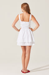 A girl is wearing a sleeveless white dress with wide straps and a fitted bodice featuring smocking details. The skirt is flared with a layered design, creating a full, gathered look. She has on red flat shoes.